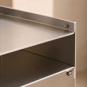 May include: A silver-colored metal shelving unit featuring two horizontal shelves with raised edges. The structure is held together by visible square-shaped metal fasteners at the corners, showcasing a smooth, brushed metallic finish throughout the piece.