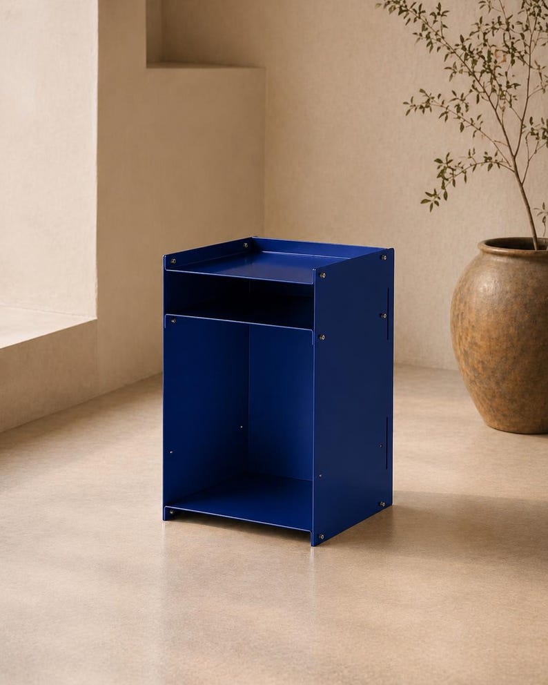 May include: A cobalt blue metal side table featuring a rectangular top surface with raised edges, a single open storage shelf below, and a larger open compartment at the base. The structure is held together by visible silver-toned metal screws.