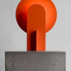 May include: Abstract sculpture featuring a vibrant orange sphere and rectangular shapes atop a gray, geometric base. The sculpture's design includes a semi-circular element and is set against a plain white background.