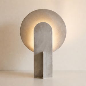 May include: A gray concrete table lamp featuring a large circular backplate and a vertical arched front panel. The design incorporates a warm, soft light that emanates from behind the circular plate, creating a glowing halo effect around the structure.