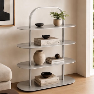 May include: A contemporary silver metal shelving unit with three oval shelves. The top shelf features a small potted plant and a black bowl. The middle shelf displays a vase, books, and a wooden sphere. The bottom shelf has books and a black bowl.