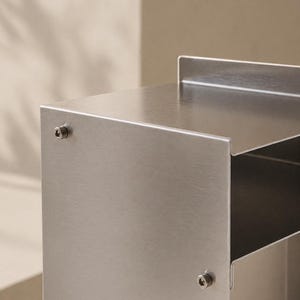 May include: A brushed stainless steel storage unit featuring a flat top surface with a raised back edge, a rectangular open compartment, and a vertical side panel secured with two visible silver-toned hexagonal bolts on the exterior surface.