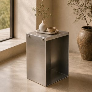 May include: A square, brushed metal side table. A small white vase with greenery, a small bowl, and a rectangular tray are on top. A large, textured, brown vase with greenery is in the background.