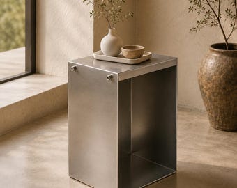 May include: A brushed metal side table with a square design. A small white vase with greenery, a small bowl, and a rectangular tray sit on top. A large, textured, brown vase with greenery is in the background.