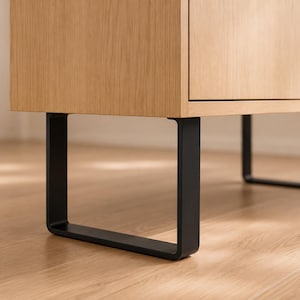 May include: Close-up of a light wood cabinet with black, square-shaped legs. The cabinet has a natural wood grain finish and a minimalist design. The legs are made of black metal.