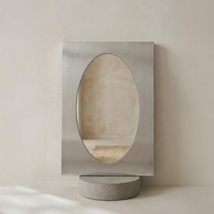May include: A rectangular, brushed metal mirror with an oval mirror in the centre. The mirror is mounted on a round, grey concrete base. The design is minimalist and modern, suitable for home decor.