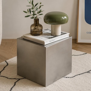 Pode incluir: Uma mesa lateral em forma de cubo de metal escovado com uma superfície lisa e reflexiva. No topo, há uma lâmpada em forma de cogumelo branca e verde-oliva, um vaso de vidro castanho com um ramo verde e um livro intitulado Bauhaus.