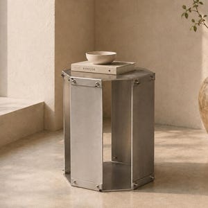 May include: A contemporary, silver metal side table with an octagonal design. A stack of books and a small, light-coloured bowl sit on the table. The table is approximately 61 cm tall. A large, textured, brown ceramic pot with a plant is in the background.