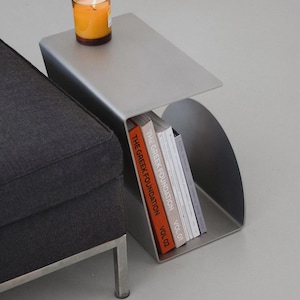 Puede incluir: Una mesa auxiliar moderna de color plateado con un diseño curvo. Una vela encendida en un tarro de cristal está sobre la mesa. La mesa contiene varios libros, incluyendo los volúmenes "The Greek Foundation".