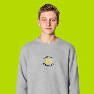 May include: A grey sweatshirt featuring the text "Caracas Es Caracas" in a yellow and black circular design. The sweatshirt is worn by a person against a bright green backdrop. The person is wearing white trousers.