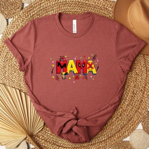 May include: A rust-colored t-shirt with the word "MAMA" in a colorful, comic book style. The letters are decorated with superhero-themed graphics, including hearts, stars, and character illustrations. The shirt is made of soft material.