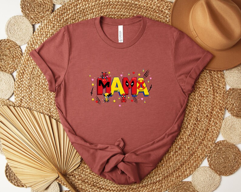May include: A rust-colored t-shirt with the word "MAMA" in a colorful, comic book style. The letters are decorated with superhero-themed graphics, including hearts, stars, and character illustrations. The shirt is made of soft material.