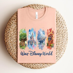 May include: A peach-colored t-shirt featuring a watercolor design of Walt Disney World landmarks. The design includes the Tree of Life, Cinderella Castle, Spaceship Earth, and the Tower of Terror, with the words "Walt Disney World" in navy blue.