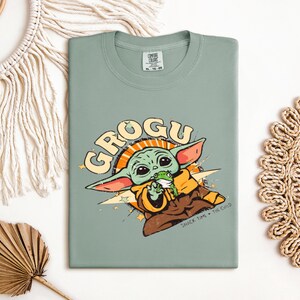 May include: Sage green t-shirt featuring a Grogu graphic. The design includes the word "GROGU" in a retro font, along with an illustration of Grogu holding a frog. The text "SNACK TIME * THE CHILD" is also included.