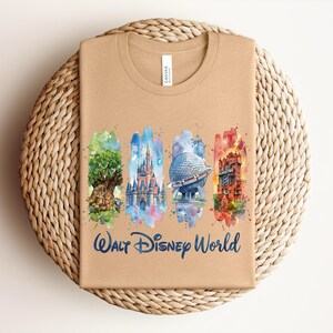 May include: Tan t-shirt featuring a watercolor design of Walt Disney World landmarks. The design includes the Tree of Life, Cinderella Castle, Spaceship Earth, and the Tower of Terror, with "Walt Disney World" written below.