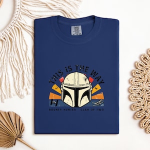 May include: A navy blue t-shirt with a graphic of a Mandalorian helmet and the phrase "THIS IS THE WAY". The design incorporates orange, yellow, and red elements, with the text "BOUNTY HUNTER CLAN OF TWO" beneath the helmet.