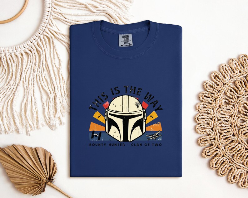 May include: A navy blue t-shirt with a graphic of a Mandalorian helmet and the phrase "THIS IS THE WAY". The design incorporates orange, yellow, and red elements, with the text "BOUNTY HUNTER CLAN OF TWO" beneath the helmet.