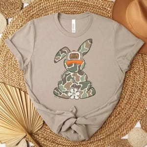 May include: A light tan t-shirt featuring a cartoon bunny wearing a camouflage pattern and a baseball cap. The bunny has a white flower on its belly. The hat says "Live Younger".