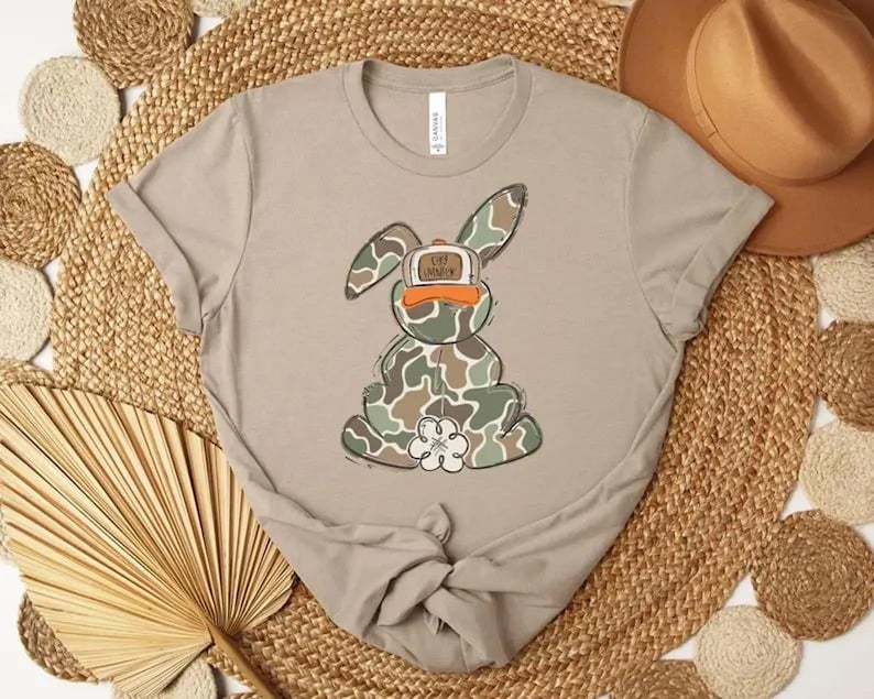 May include: A light tan t-shirt featuring a cartoon bunny wearing a camouflage pattern and a baseball cap. The bunny has a white flower on its belly. The hat says "Live Younger".