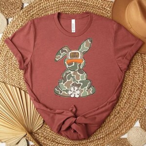 May include: A maroon t-shirt featuring a cartoon bunny with a camouflage pattern. The bunny has a duck bill and a flower on its chest. The text on the bunny's hat reads "OFF DUTY".