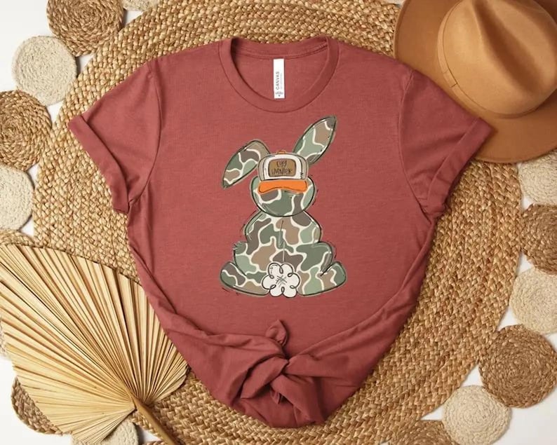 May include: A maroon t-shirt featuring a cartoon bunny with a camouflage pattern. The bunny has a duck bill and a flower on its chest. The text on the bunny's hat reads "OFF DUTY".