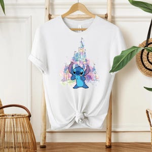 Pu&ograve; includere: T-shirt bianco con un personaggio dei cartoni animati blu e un castello sullo sfondo. Il castello &egrave; raffigurato in stile acquerello con tonalit&agrave; rosa, viola e blu. La maglietta &egrave; annodata in vita.