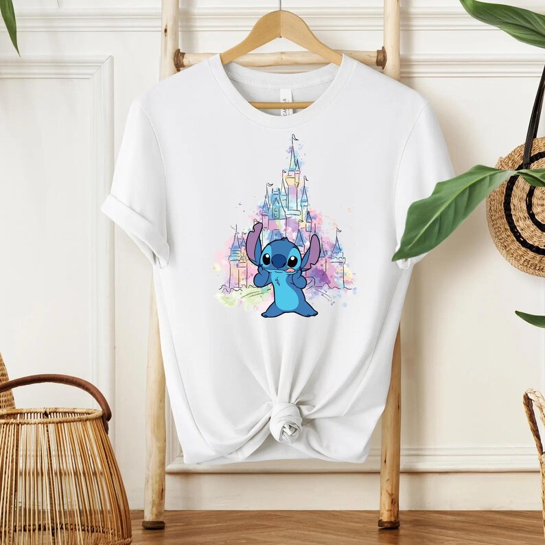 Pu&ograve; includere: T-shirt bianco con un personaggio dei cartoni animati blu e un castello sullo sfondo. Il castello &egrave; raffigurato in stile acquerello con tonalit&agrave; rosa, viola e blu. La maglietta &egrave; annodata in vita.