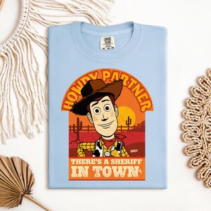 K&ouml;nnte beinhalten: Hellblaues T-Shirt mit einer Cartoon-Illustration von Woody aus Toy Story. Der Aufdruck zeigt den Text "HOWDY PARTNER" und "THERE'S A SHERIFF IN TOWN". Das Design ist vor einer W&uuml;sten-Sonnenuntergangs-Kulisse platziert.