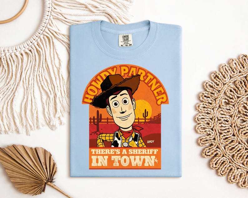 K&ouml;nnte beinhalten: Hellblaues T-Shirt mit einer Cartoon-Illustration von Woody aus Toy Story. Der Aufdruck zeigt den Text "HOWDY PARTNER" und "THERE'S A SHERIFF IN TOWN". Das Design ist vor einer W&uuml;sten-Sonnenuntergangs-Kulisse platziert.