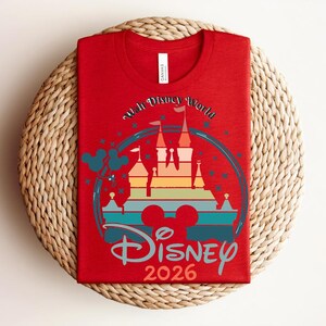 Op de afbeelding: Een rood t-shirt met een kleurrijke afbeelding van een kasteel, de tekst "Walt Disney World" en "Disney 2026". Het ontwerp bevat Mickey Mouse oren en een regenboogkleurige kasteelsilhouet. Voor Disney-liefhebbers.