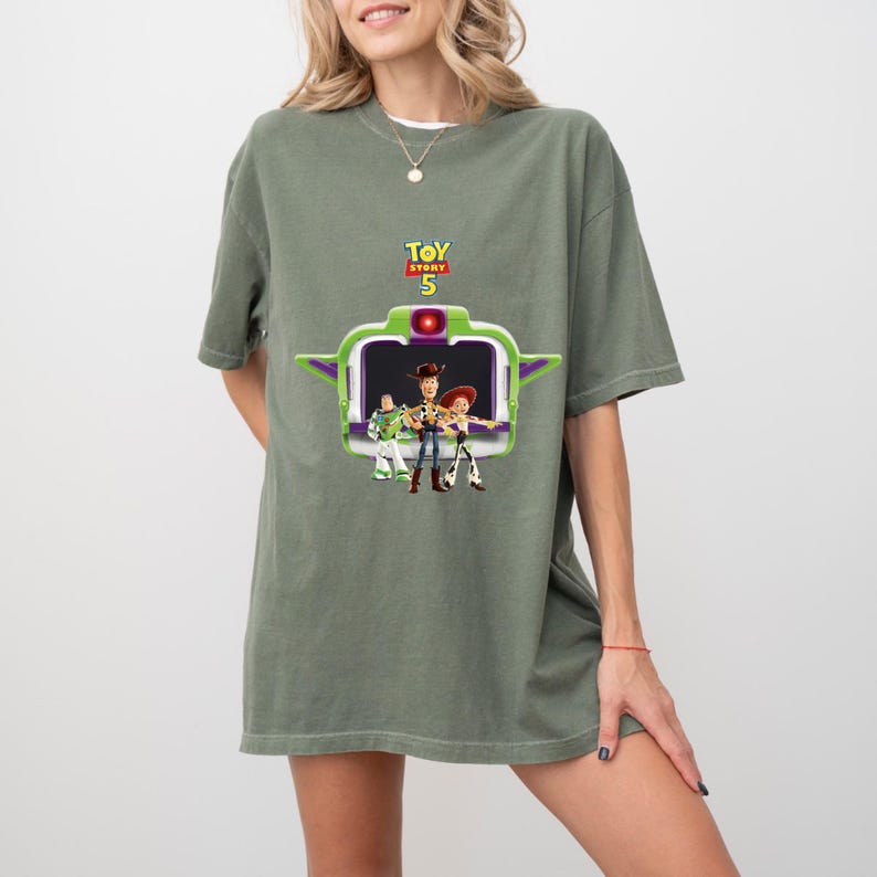 K&ouml;nnte beinhalten: Ein olivgr&uuml;nes T-Shirt mit einer Grafik aus dem Film Toy Story 5. Die Grafik zeigt Buzz Lightyear, Woody und Jessie in einem gr&uuml;n-violetten raumschiffartigen Rahmen. Der Text "TOY STORY 5" befindet sich &uuml;ber der Grafik.