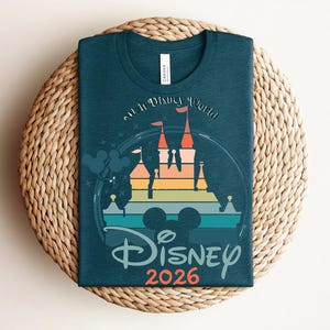 Op de afbeelding: Tealkleurig T-shirt met een kleurrijke afbeelding van een kasteel en de tekst "Disney 2026". Het ontwerp bevat een regenboog aan kleuren en de iconische Mickey Mouse oren. Het shirt ligt op een geweven, ronde placemat.
