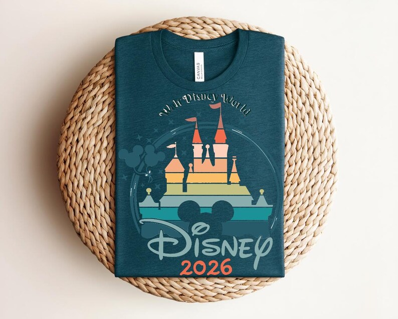 Op de afbeelding: Tealkleurig T-shirt met een kleurrijke afbeelding van een kasteel en de tekst "Disney 2026". Het ontwerp bevat een regenboog aan kleuren en de iconische Mickey Mouse oren. Het shirt ligt op een geweven, ronde placemat.