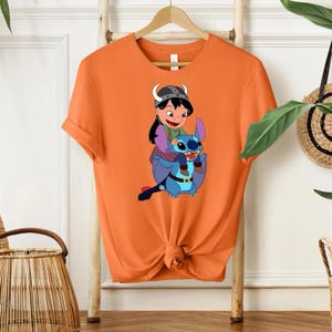 Może przedstawiać: Pomarańczowa koszulka z kresk&oacute;wkową ilustracją dziewczynki jadącej na niebieskim stworzeniu przypominającym kosmitę. Dziewczynka ma na sobie hełm, a stw&oacute;r duże uszy. Projekt jest wyśrodkowany na koszulce.