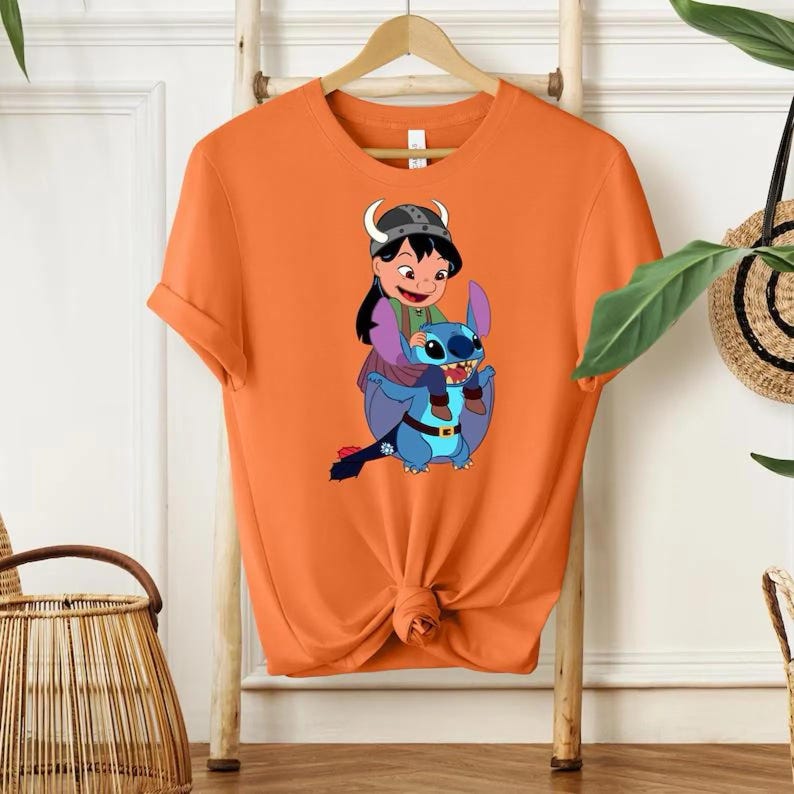 Może przedstawiać: Pomarańczowa koszulka z kresk&oacute;wkową ilustracją dziewczynki jadącej na niebieskim stworzeniu przypominającym kosmitę. Dziewczynka ma na sobie hełm, a stw&oacute;r duże uszy. Projekt jest wyśrodkowany na koszulce.