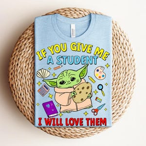 May include: Light blue t-shirt with a graphic of a green alien holding a cookie, surrounded by school supplies. Text reads "IF YOU GIVE ME A STUDENT I WILL LOVE THEM" in yellow and red.