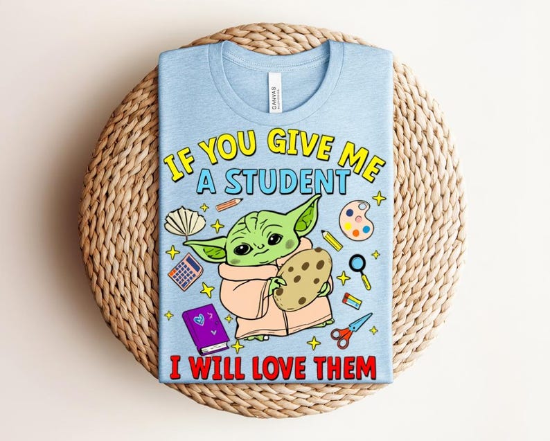 May include: Light blue t-shirt with a graphic of a green alien holding a cookie, surrounded by school supplies. Text reads "IF YOU GIVE ME A STUDENT I WILL LOVE THEM" in yellow and red.