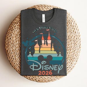 Op de afbeelding: Donkergrijs t-shirt met een kleurrijke afbeelding van een kasteel, de tekst "Walt Disney World" en "Disney 2026". Het ontwerp bevat een regenboog kasteel silhouet en Mickey Mouse oren.