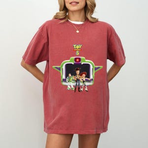 K&ouml;nnte beinhalten: Ein rotes T-Shirt mit einer Grafik aus dem Film Toy Story 5. Die Grafik zeigt Buzz Lightyear, Woody und Jessie. Das T-Shirt hat einen Rundhalsausschnitt und kurze &Auml;rmel.