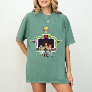 K&ouml;nnte beinhalten: Ein gr&uuml;nes T-Shirt mit einem Toy Story 5-Grafik. Das Design zeigt Buzz Lightyear, Woody und Jessie in einem gr&uuml;n-violetten raumschiffartigen Rahmen. Der Text "Toy Story 5" ist in Gelb und Rot &uuml;ber der Grafik dargestellt.