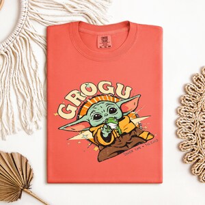May include: Coral-colored t-shirt featuring a graphic of Grogu, the character from The Mandalorian. The design includes Grogu holding a frog, with the word "GROGU" in a retro font. The phrase "SNACK TIME - THE CHILD" is also present.