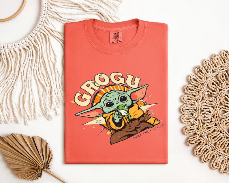 May include: Coral-colored t-shirt featuring a graphic of Grogu, the character from The Mandalorian. The design includes Grogu holding a frog, with the word "GROGU" in a retro font. The phrase "SNACK TIME - THE CHILD" is also present.