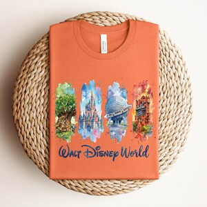 May include: Orange t-shirt featuring watercolor illustrations of iconic Walt Disney World landmarks. The design includes images of the Tree of Life, Cinderella Castle, Spaceship Earth, and the Tower of Terror, with "Walt Disney World" written below.