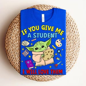 May include: Royal blue t-shirt with the text "IF YOU GIVE ME A STUDENT I WILL LOVE THEM". The graphic features a green alien-like character holding a cookie, surrounded by school-themed illustrations. The shirt is displayed on a woven mat.