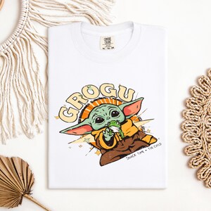 May include: White t-shirt featuring a Grogu graphic. The design includes the text "GROGU" in yellow and orange, with an illustration of Grogu holding a frog. The phrase "SNACK TIME - THE CHILD" is also present.