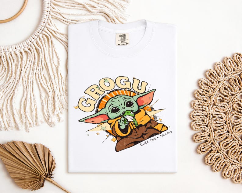 May include: White t-shirt featuring a Grogu graphic. The design includes the text "GROGU" in yellow and orange, with an illustration of Grogu holding a frog. The phrase "SNACK TIME - THE CHILD" is also present.
