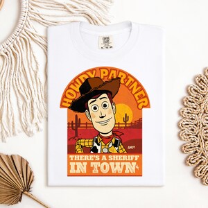 K&ouml;nnte beinhalten: Wei&szlig;es T-Shirt mit einer Cartoon-Illustration eines Cowboys. Das Design enth&auml;lt den Text "HOWDY PARTNER" und "THERE'S A SHERIFF IN TOWN" vor einem Sonnenuntergangshintergrund. Der Name der Figur, "ANDY", ist ebenfalls zu sehen.