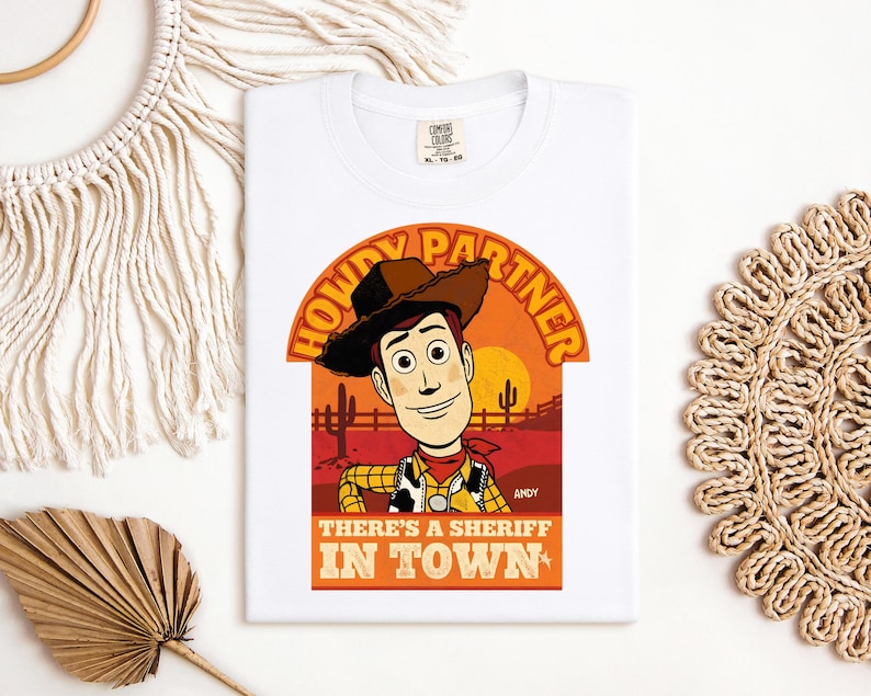 K&ouml;nnte beinhalten: Wei&szlig;es T-Shirt mit einer Cartoon-Illustration eines Cowboys. Das Design enth&auml;lt den Text "HOWDY PARTNER" und "THERE'S A SHERIFF IN TOWN" vor einem Sonnenuntergangshintergrund. Der Name der Figur, "ANDY", ist ebenfalls zu sehen.