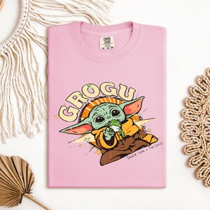 May include: A light pink t-shirt featuring a cartoon graphic of Grogu, also known as Baby Yoda, from The Mandalorian. The graphic includes the word "GROGU" in a retro font, along with the phrase "SNACK TIME + THE CHILD". The shirt is made of a soft material.