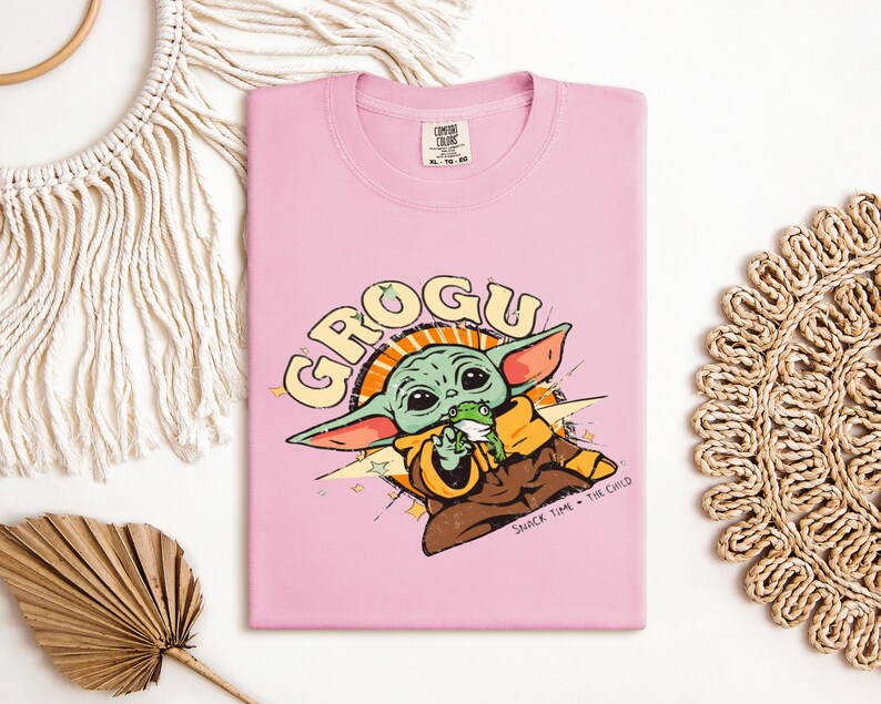 May include: A light pink t-shirt featuring a cartoon graphic of Grogu, also known as Baby Yoda, from The Mandalorian. The graphic includes the word "GROGU" in a retro font, along with the phrase "SNACK TIME + THE CHILD". The shirt is made of a soft material.